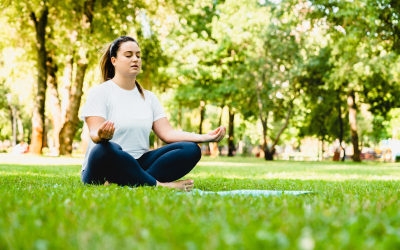 Zen-like caucasian calm young woman plus size fat athlete meditating on yoga class, training in fitness outfit outdoors in city park